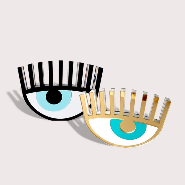 Studio photo of two plexiglass evil eye table charms (black and gold) on soft neutral background – So Cute by Dimi.