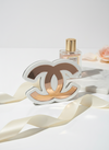 Επιτραπέζιο γούρι Double C από λευκό και ροζ χρυσό καθρέφτη, τοποθετημένο σε minimal λευκή επιφάνεια με άρωμα Chanel στο φόντο.