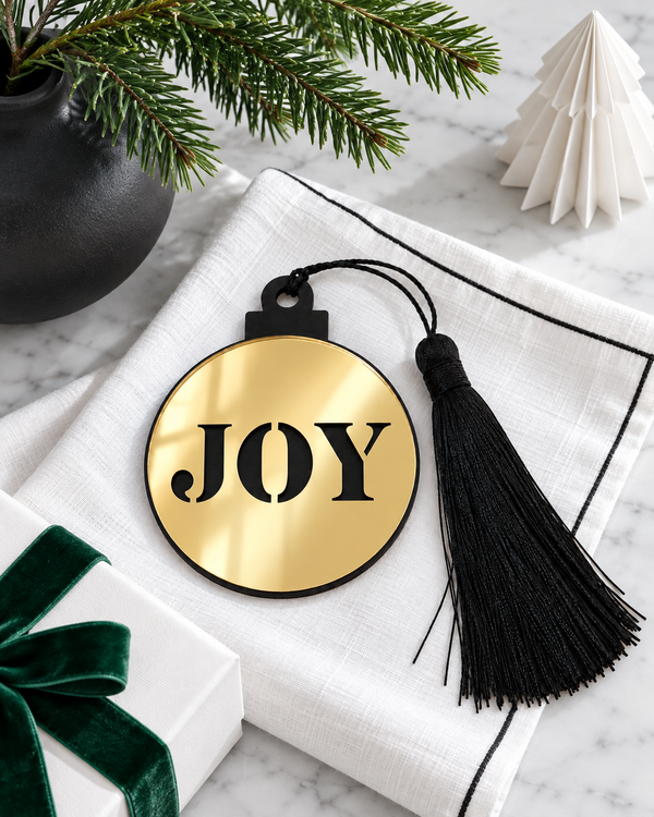 Γούρι μπάλα JOY με χρυσό καθρέφτη και μαύρη φούντα πάνω σε λευκό ύφασμα, με κλαδί έλατου, χάρτινο χριστουγεννιάτικο δέντρο και δώρο με πράσινη κορδέλα – So Cute Cut.