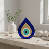 Blue and gold plexiglass teardrop evil eye ornament displayed indoors with olive branch vase.
